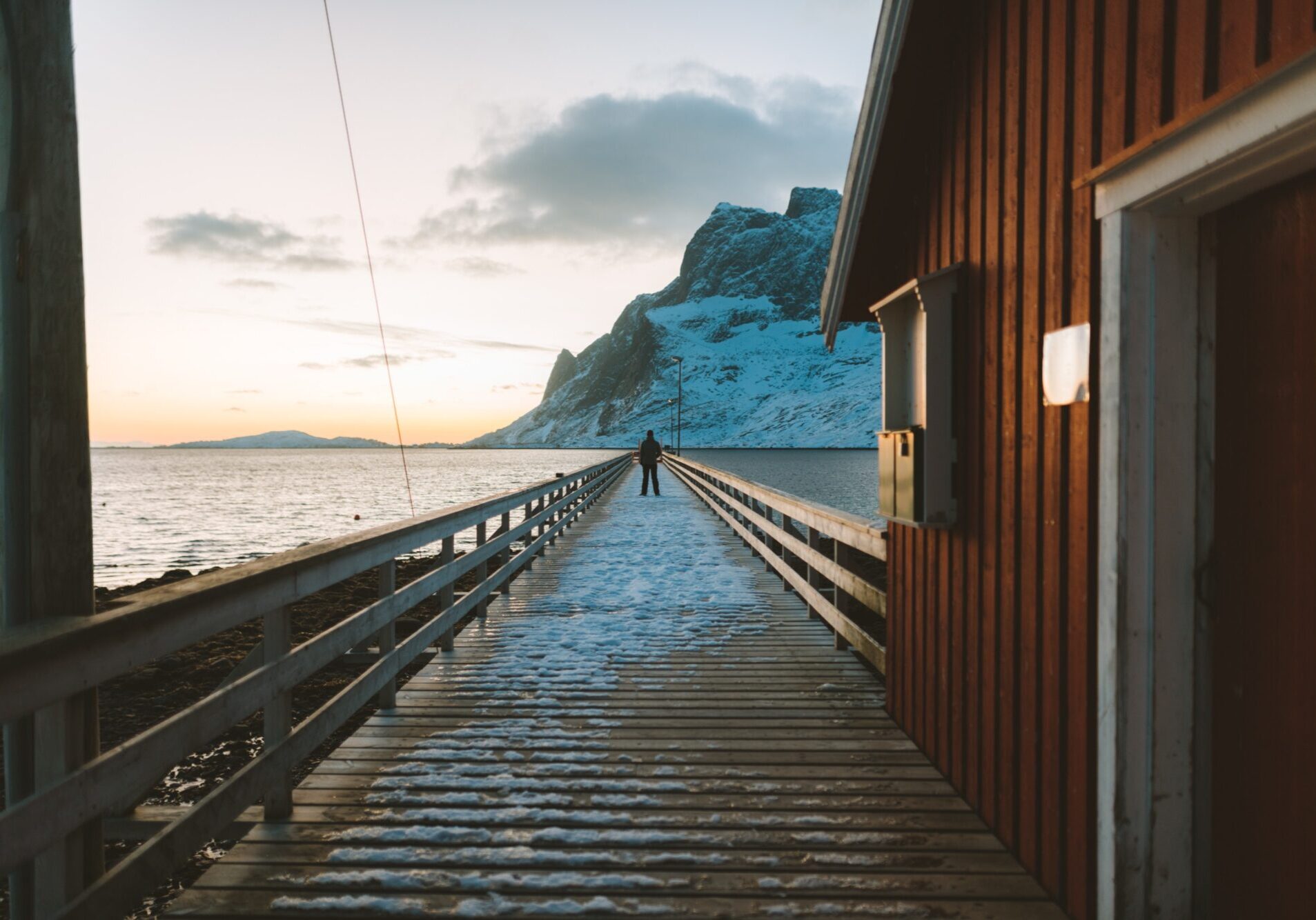 En brygge ved siden a et rødt hus, hvor en person står midt på bryggen i det fjerne